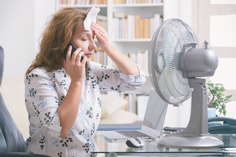 Woman using fan to cool off at desk due to her air conditioner breaking down.