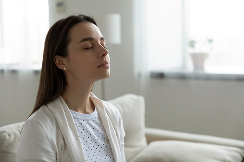 A young woman with long brown hair sits with her eyes closed and a calm expression, seemingly practicing deep breathing, meditating, or enjoying fresh indoor air.