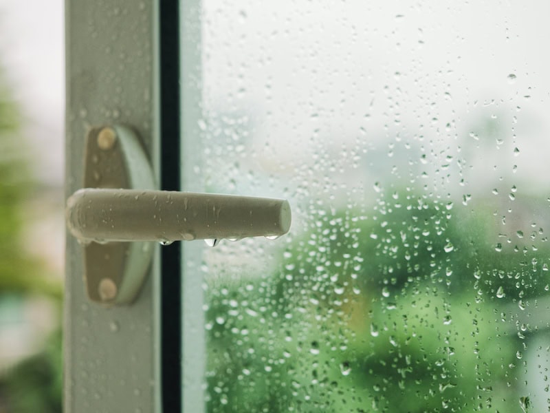 A close-up shows a white or light grey window handle covered in water droplets during a rainstorm.
